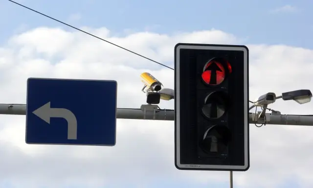 Rejestrator przejazdu na czerwonym świetle, widoczny na tle nieba, wraz z sygnalizacją świetlną i znakiem nakazu skrętu w lewo.