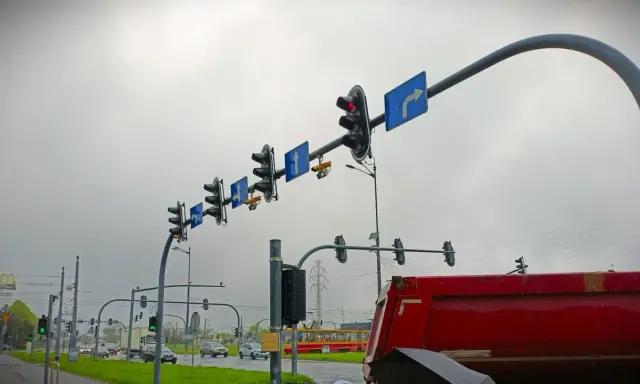 Na skrzyżowaniu wiszą sygnalizatory świetlne i znaki kierunkowe. Widać żółty tramwaj i czerwony wywrotkę. Gdzie są kamery rejestrujące przejazd na czerwonym świetle?