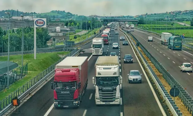 Ciężarówki na autostradzie, jedna czerwona Scania wyprzedza białą. W tle inne pojazdy i krajobraz z pagórkami.