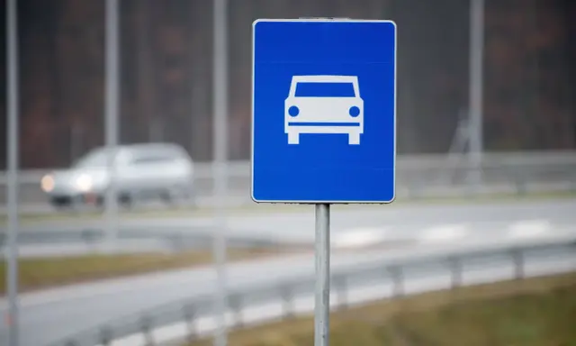 Znak drogowy szybkiego ruchu z symbolem samochodu. W tle widać rozmazany pojazd na drodze.