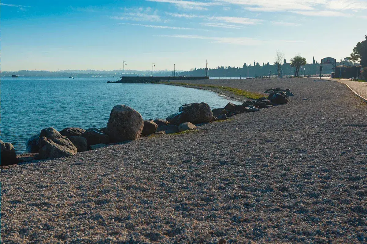 Kamienista plaża nad jeziorem, idealna na spacer po Peschiera del Garda. Widok na spokojną wodę i zarys miasta.