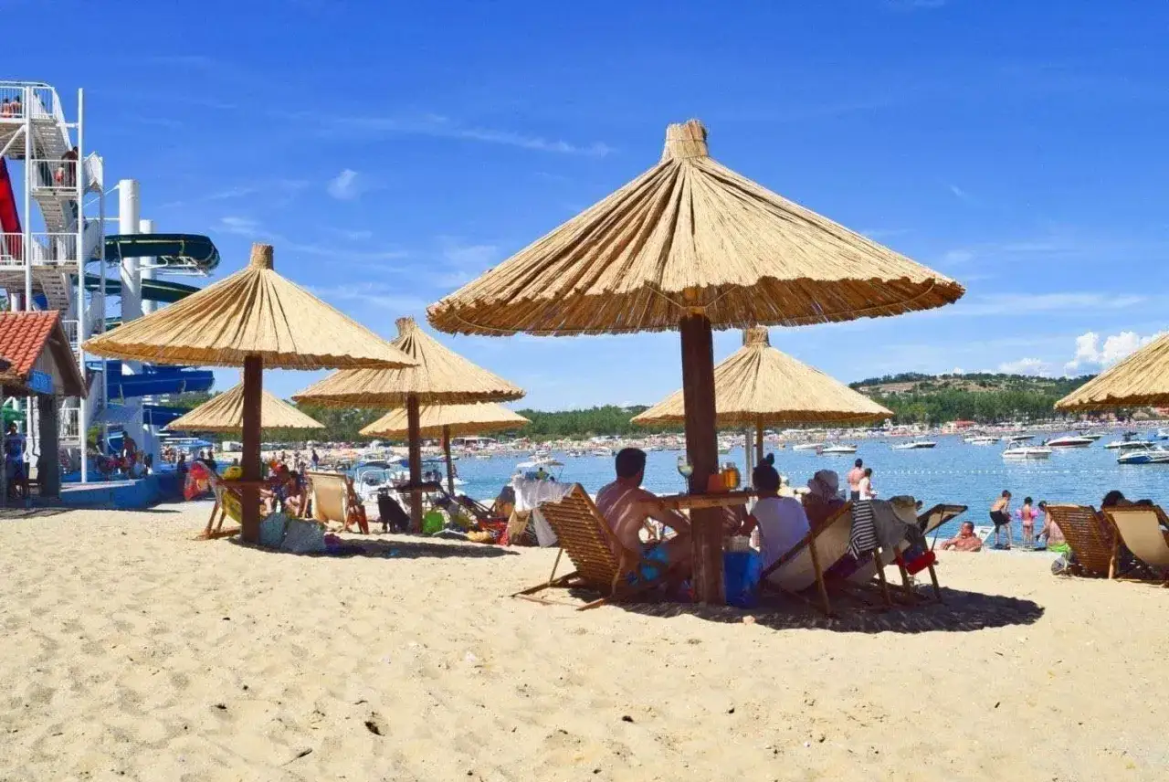 Słoneczna plaża na wyspie Rab, z ludźmi odpoczywającymi pod strzechami, w tle woda z łodziami i zjeżdżalnie wodne.