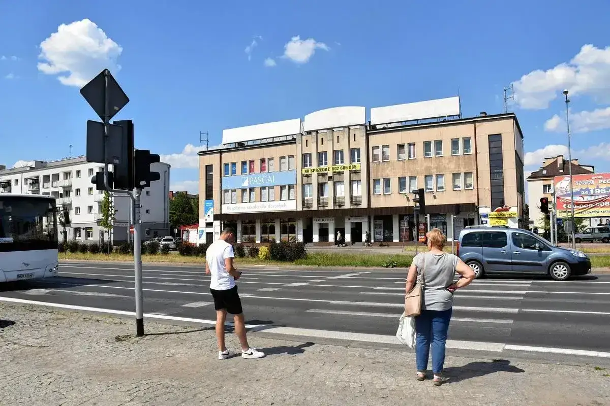 Ludzie czekają na zielone światło, by przejść przez pasy. Zastanawiają się, jak dojechać do Płocka, patrząc na drogę i budynki.