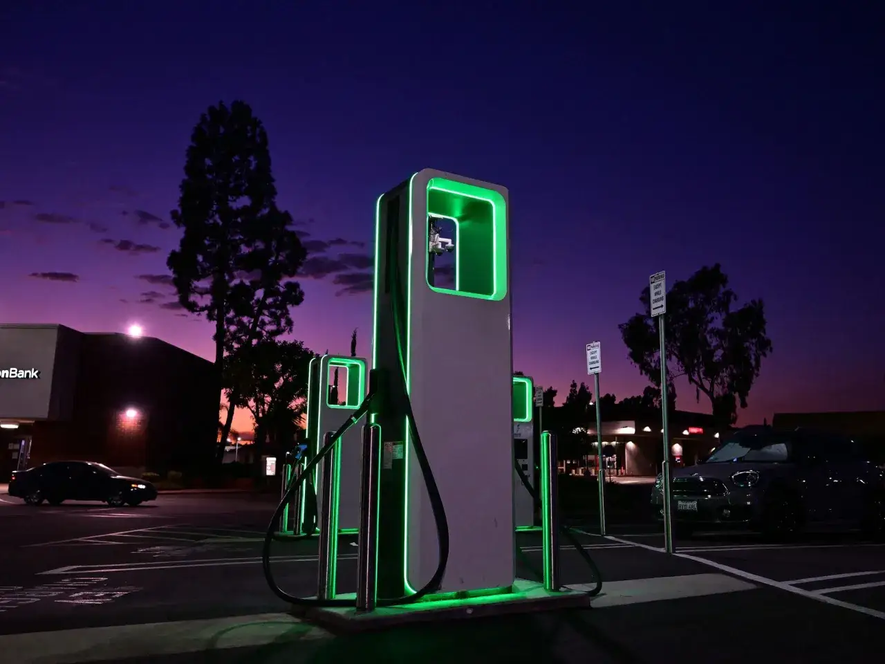 Stacja ładowania samochodów elektrycznych nocą. Zielone światła podkreślają nowoczesne ładowarki, a fioletowe niebo zapowiada długi zasięg samochodu elektrycznego.