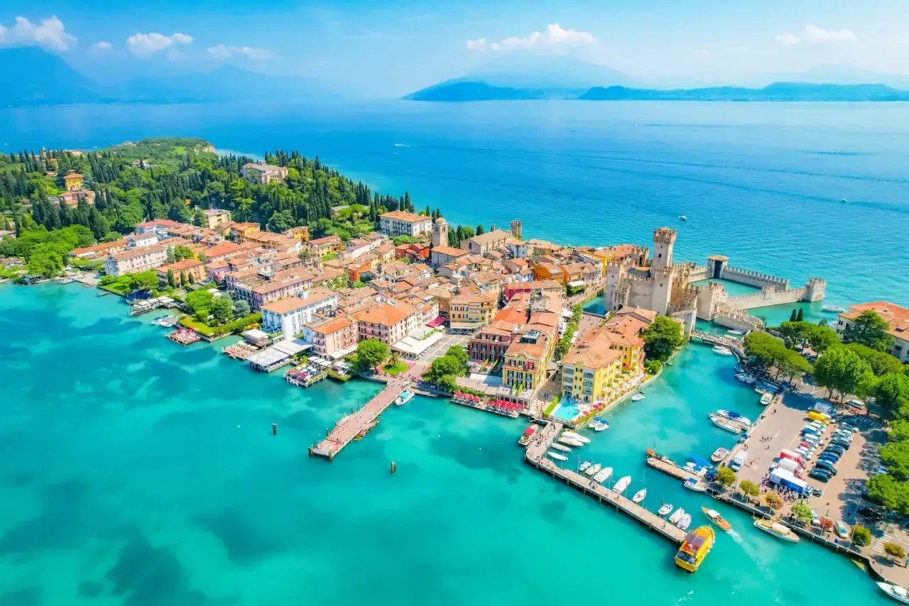 Malownicze jezioro Garda z plażami, zamkiem i kolorowymi budynkami. Woda kusi turkusowym odcieniem, a łodzie kołyszą się przy pomostach.