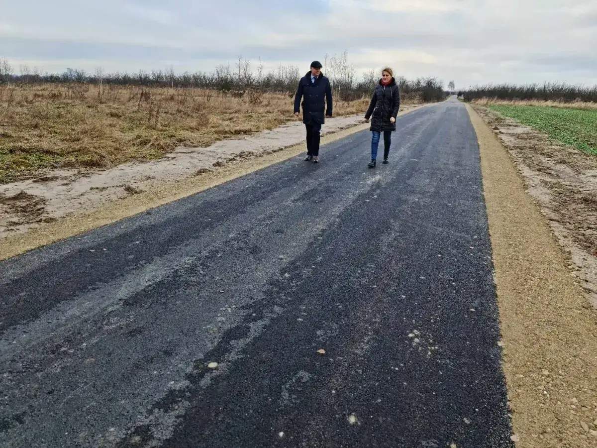 Para idzie nową drogą lokalną, otoczoną polami i krzewami.