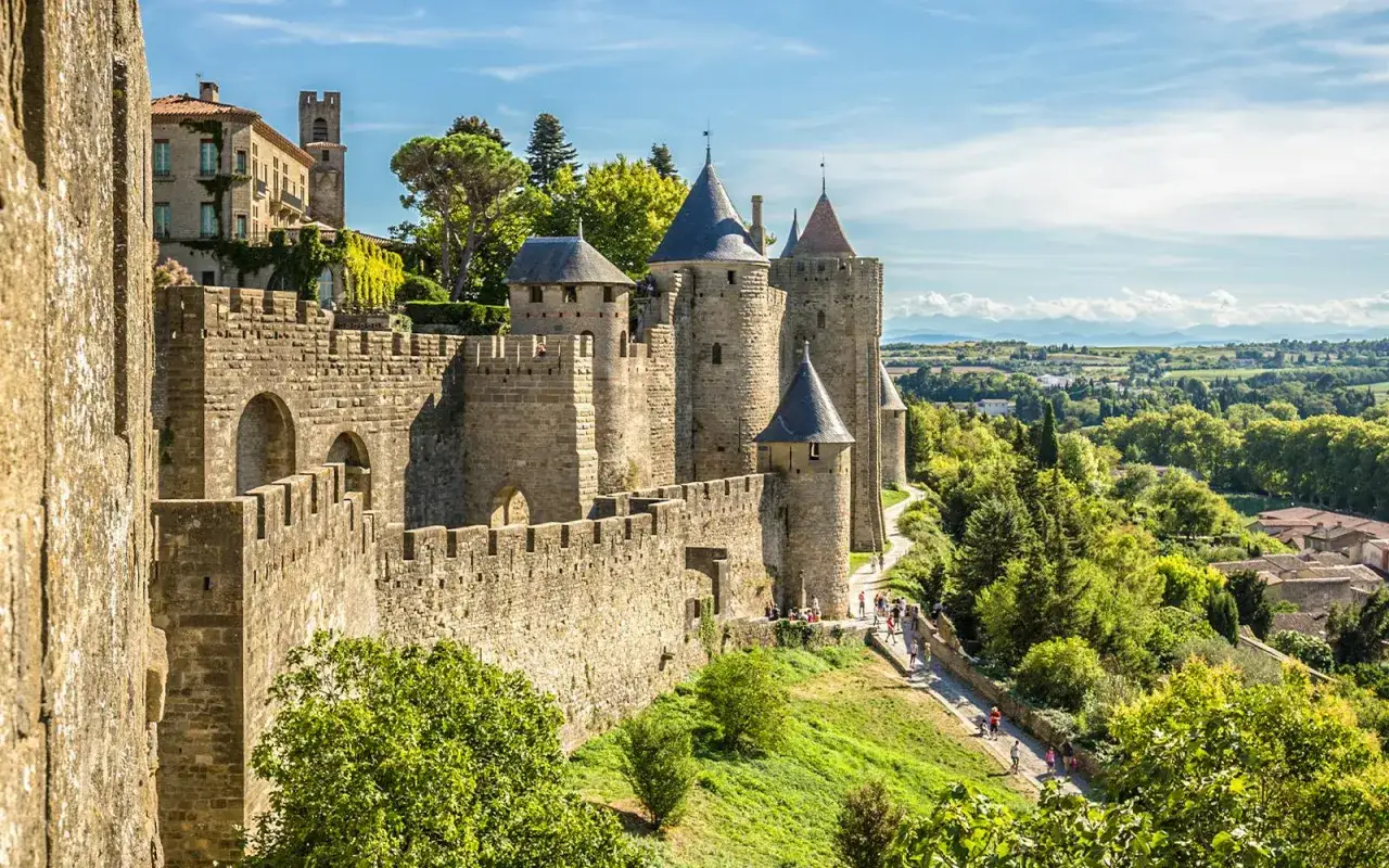Kamienne mury i wieże średniowiecznego miasta Carcassonne we Francji, otoczone zielonymi wzgórzami i błękitnym niebem.