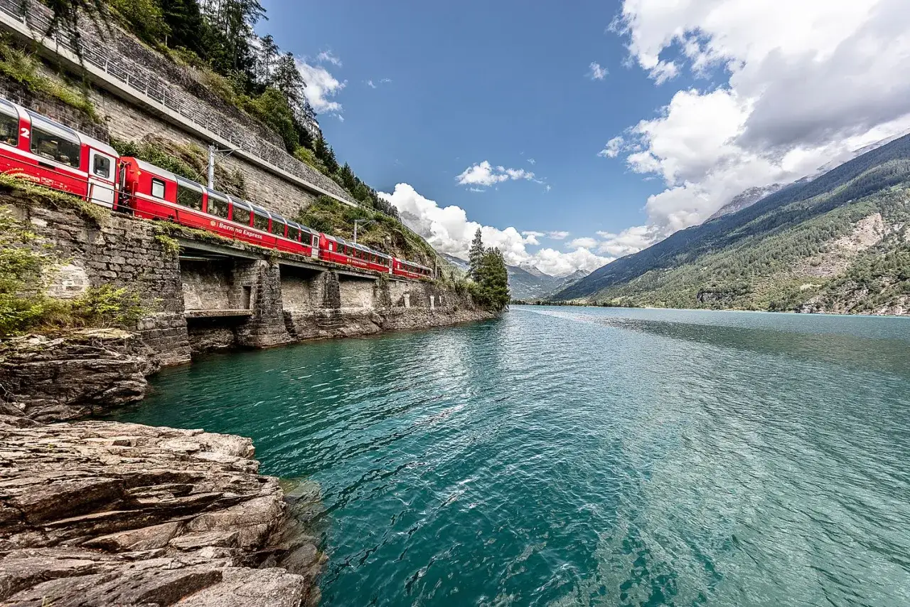 Czerwony pociąg Bernina Express jedzie wzdłuż turkusowego jeziora w Szwajcarii latem. Góry porośnięte lasem i skaliste brzegi.