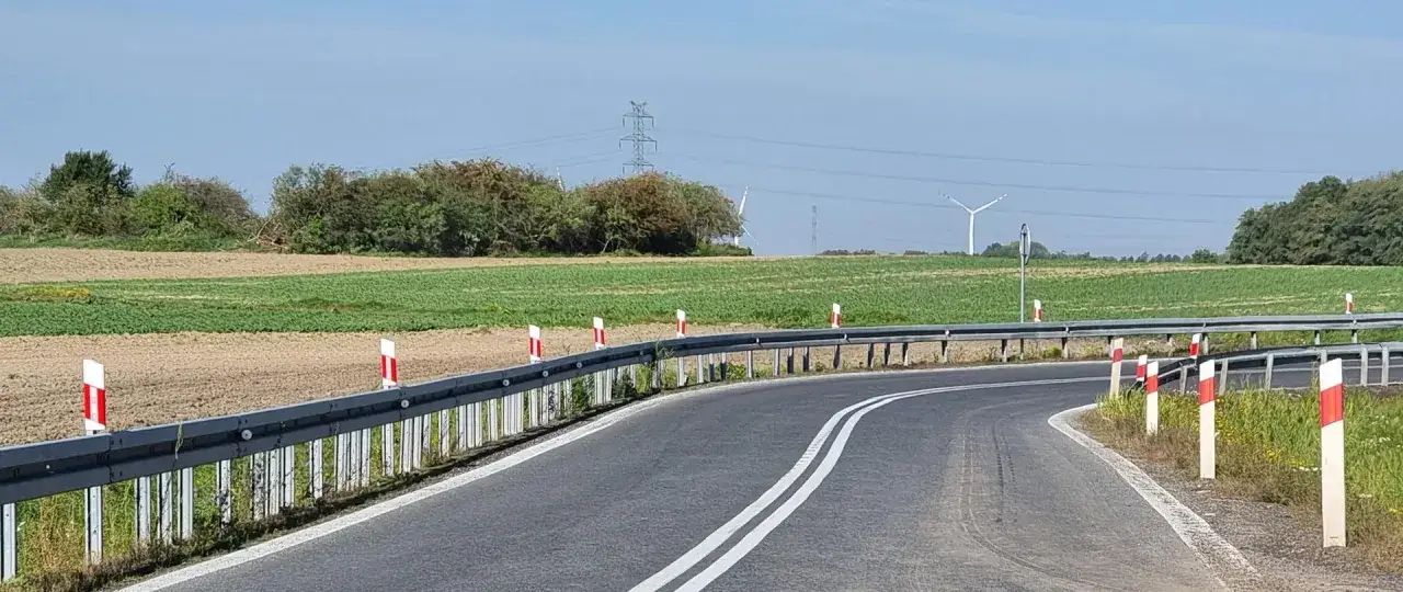 Słupki na autostradzie wyznaczają zakręt drogi wśród pól. W tle widać wiatraki i linie energetyczne.