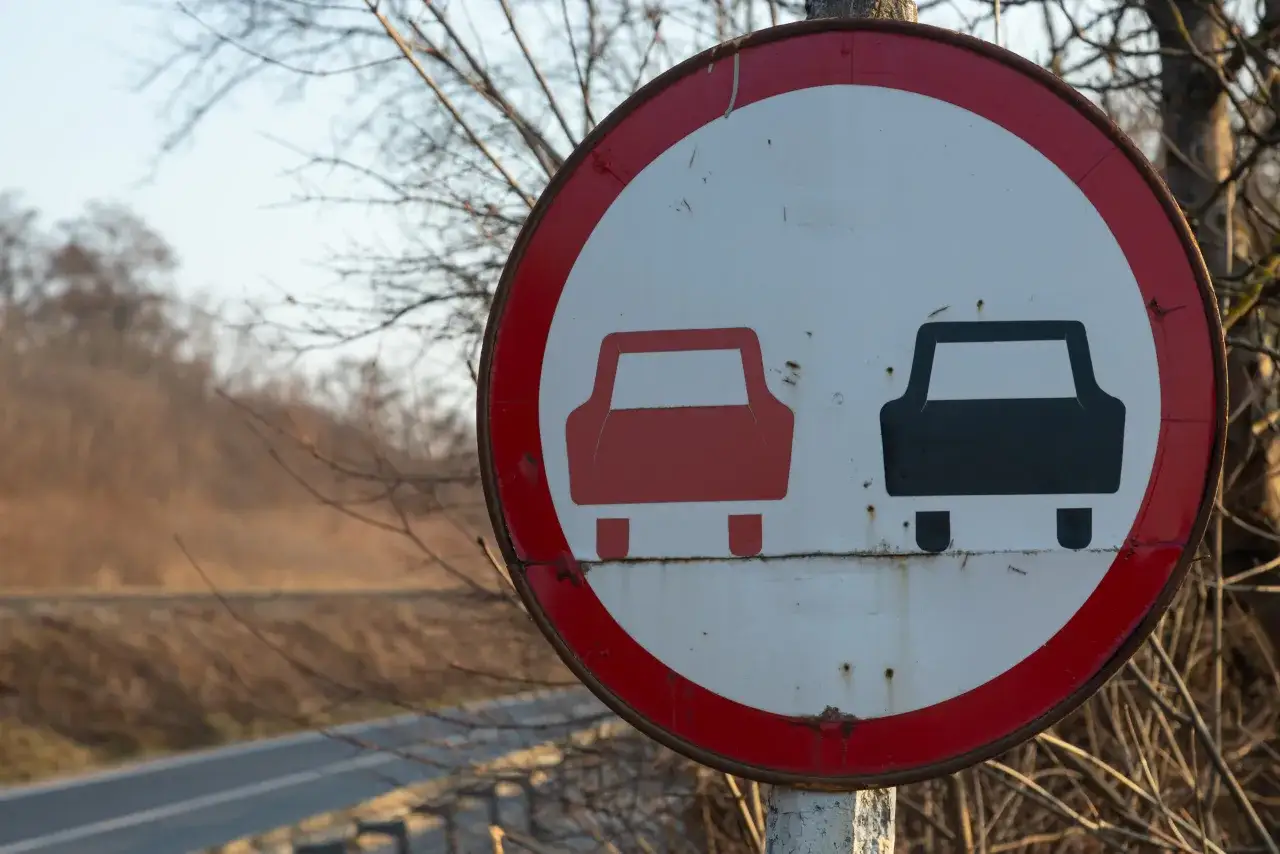 Znak zakazu wyprzedzania. Czerwony samochód zablokowany przez czarny, ostrzega przed niebezpiecznym manewrem wyprzedzania.