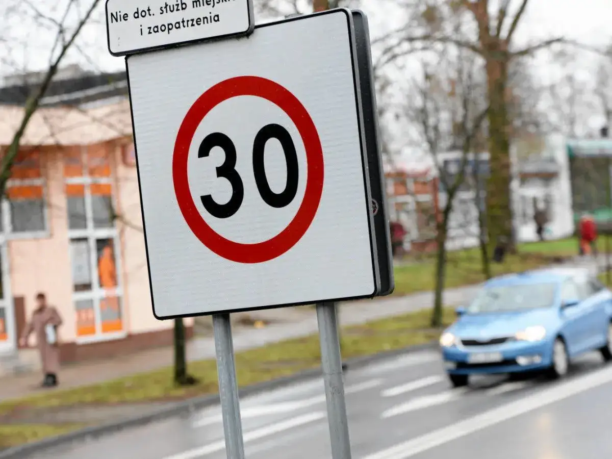 Znak ograniczenia prędkości do 30 km/h. Na drodze ekspresowej, w tle budynek i niebieski samochód.