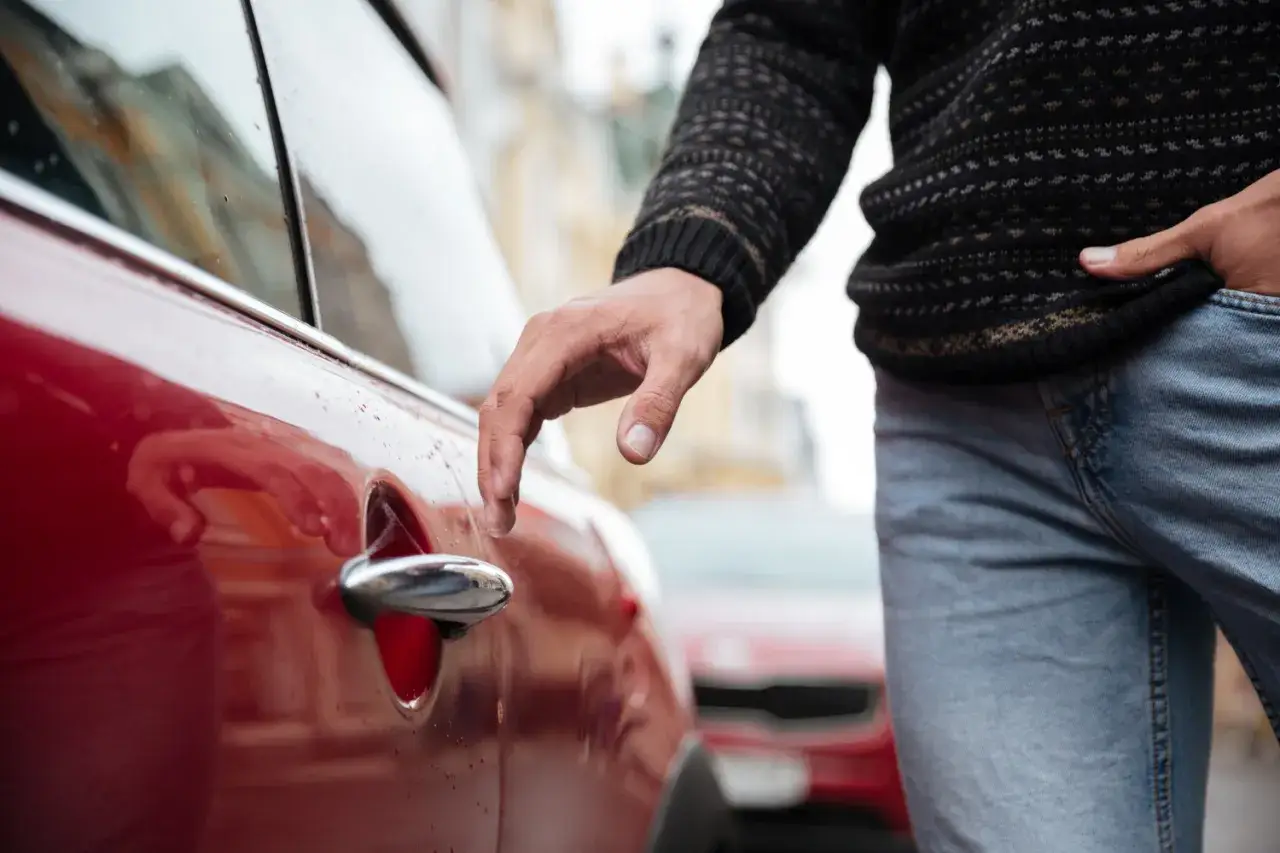 Zatrzaśnięte kluczyki w samochodzie? Mężczyzna pr&oacute;buje otworzyć czerwone auto, szukając rozwiązania.