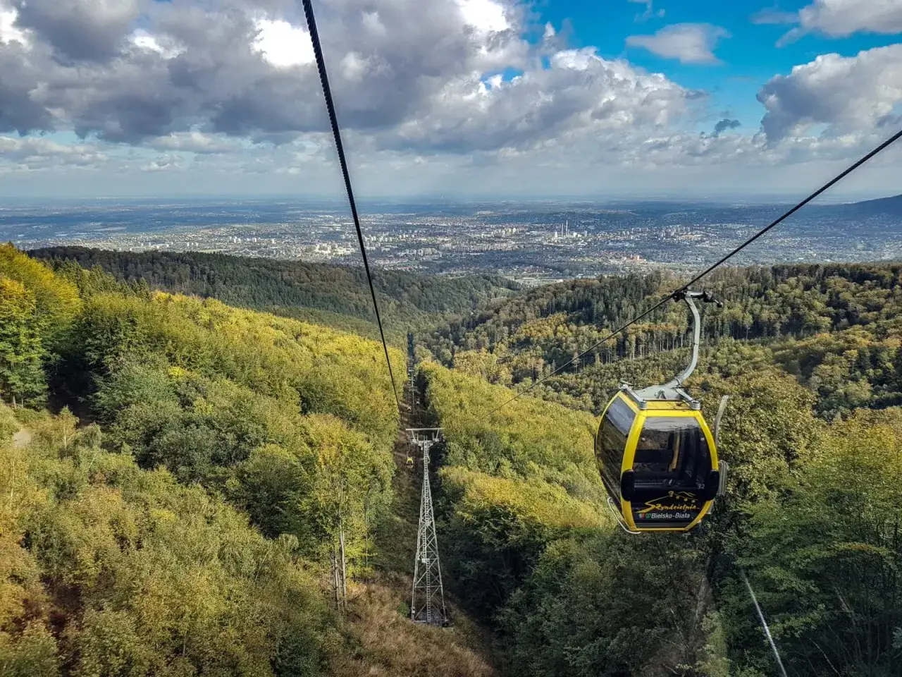 Kolejka linowa wjeżdża na górę, oferując widok na Bielsko-Białą i Szczyrk. Jesienne lasy otaczają trasę.