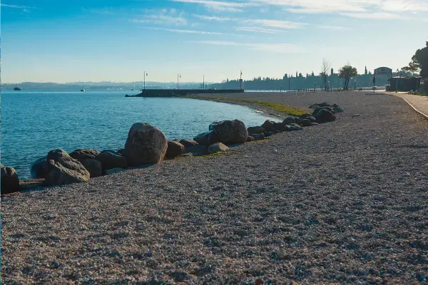 Kamienista plaża nad jeziorem, idealna na spacer po Peschiera del Garda. Widok na spokojną wodę i zarys miasta.