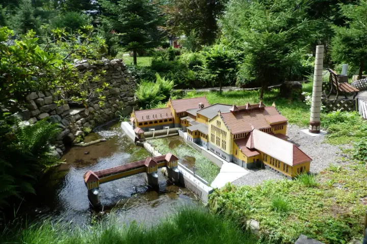 Miniaturowa zabudowa z mostkiem i rzeką, jak w parkach miniatur w Polsce. W tle drzewa i kamienny murek.